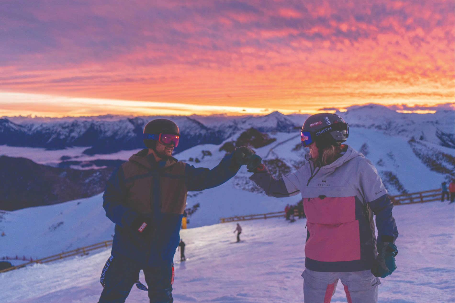 Two skiers fist bumping on a snowy mountain slope at sunset with vibrant orange and purple sky.