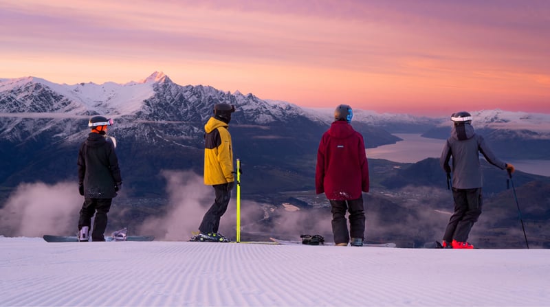 Group of skiers and snowboarders looking out over mountain and lake views at sunset